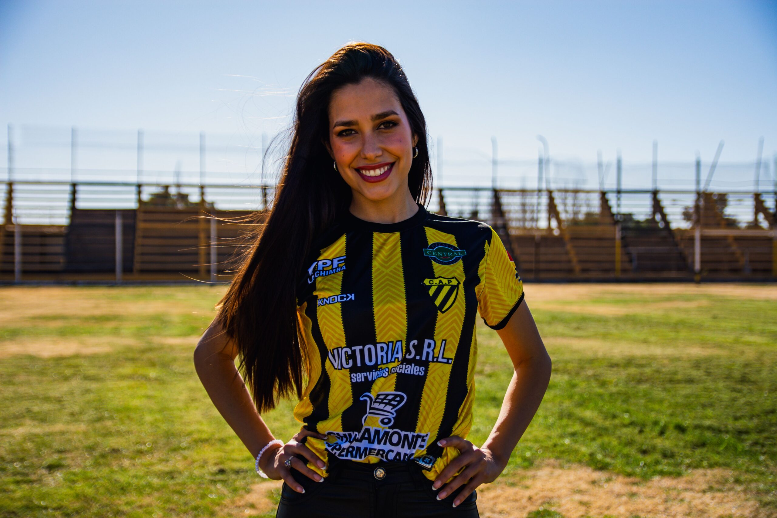 Woman in a black and yellow sports jersey standing on a sunny football field, smiling confidently.